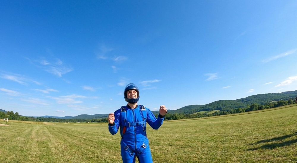 Un jeune homme en combinaison de parachutisme bleue et casque se tient dans un champ, regardant le ciel.