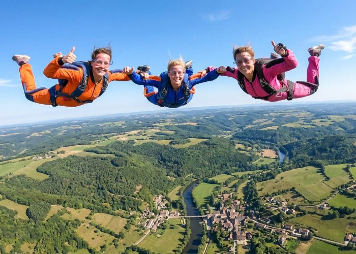 Trois femmes en combinaisons colorées sautent en parachute au-dessus d'un paysage verdoyant et d'un village.