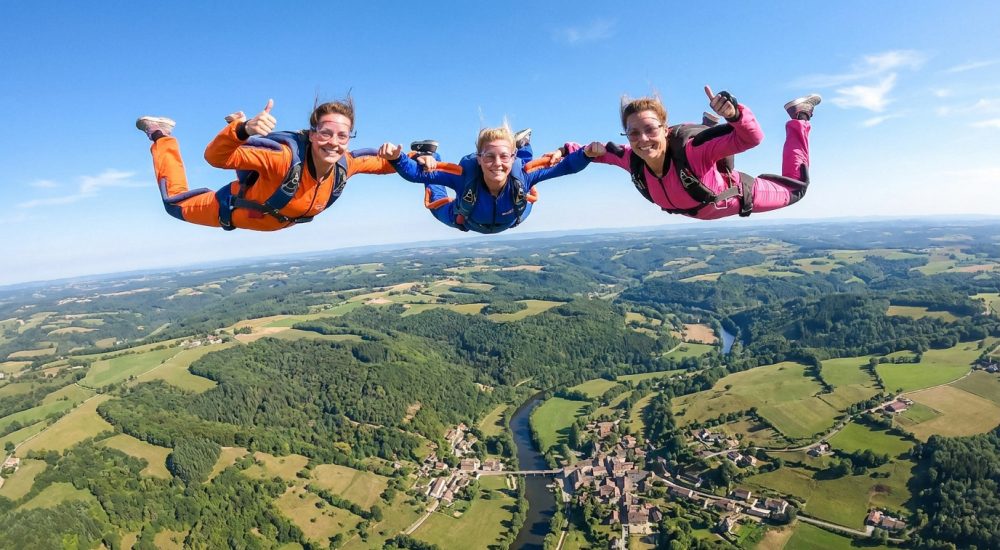 Trois femmes en combinaisons colorées sautent en parachute au-dessus d'un paysage verdoyant et d'un village.