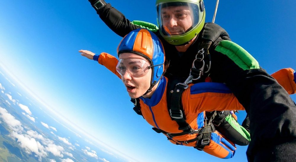 Une femme avec un moniteur en plein saut en parachute tandem, bras écartés devant un ciel bleu azur.