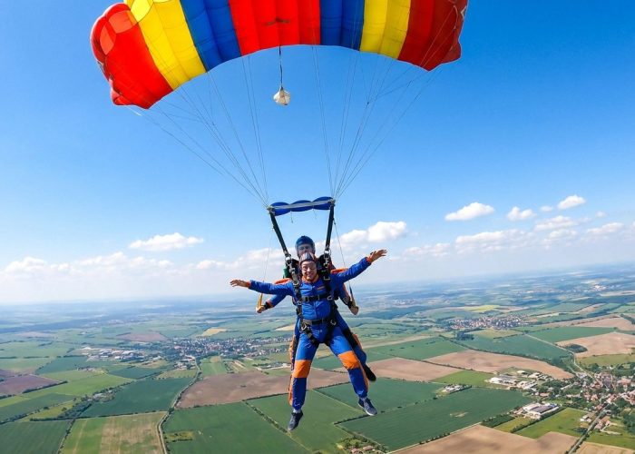 Un homme en tenue de parachutisme effectue un saut en tandem sous un grand parachute coloré au-dessus d'un paysage rural.