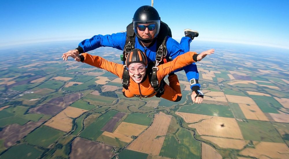 Une femme et son instructeur en chute libre au-dessus d'un paysage de champs colorés.