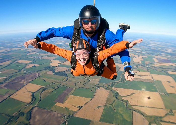 Une femme et son instructeur en chute libre au-dessus d'un paysage de champs colorés.
