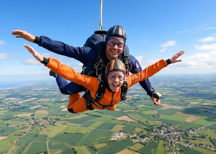 Une femme en combinaison orange effectue un saut en parachute en tandem avec un moniteur au-dessus d'une campagne.